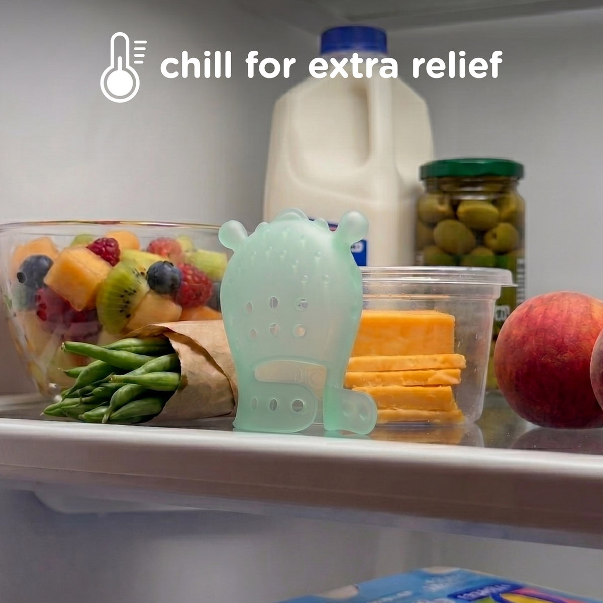 A mint green silicone refrigerator ice melt holder shaped like a small bear or creature, shown on a refrigerator shelf with food items and a milk jug in the background. White text at the top reads 'chill for extra relief'.